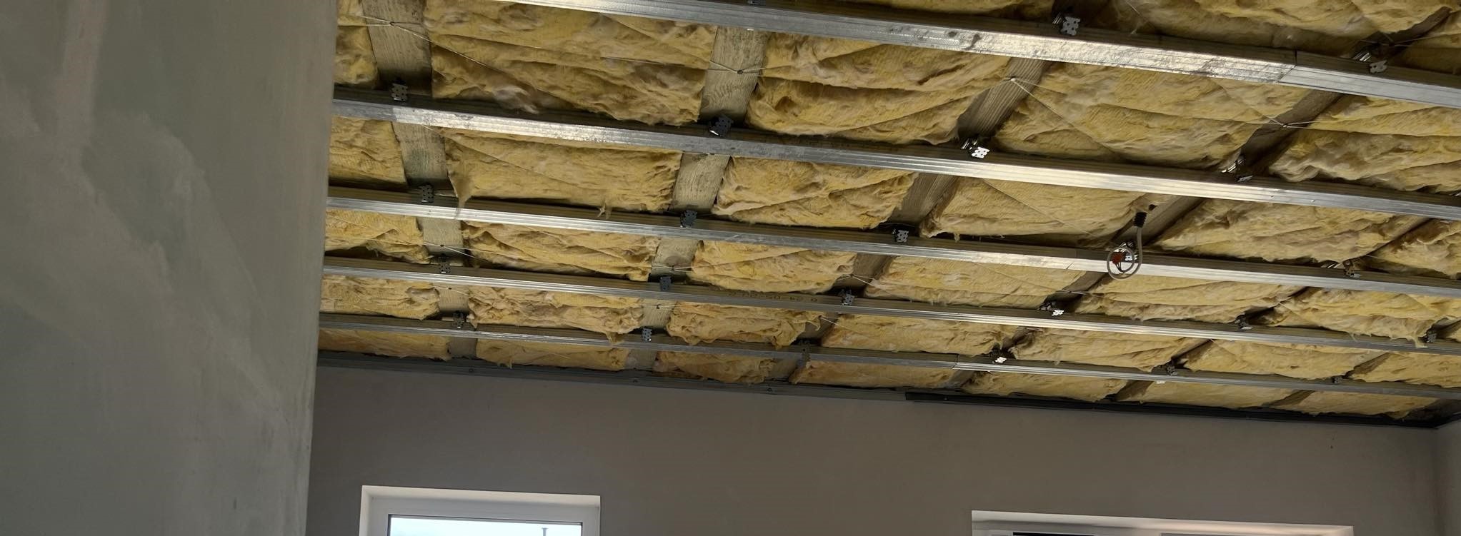 Montaż izolacji z wełny mineralnej w metalowym stelażu sufitu podwieszanego w Grodzisku Mazowieckim, widoczna szpachlowana ściana i fragment okna.