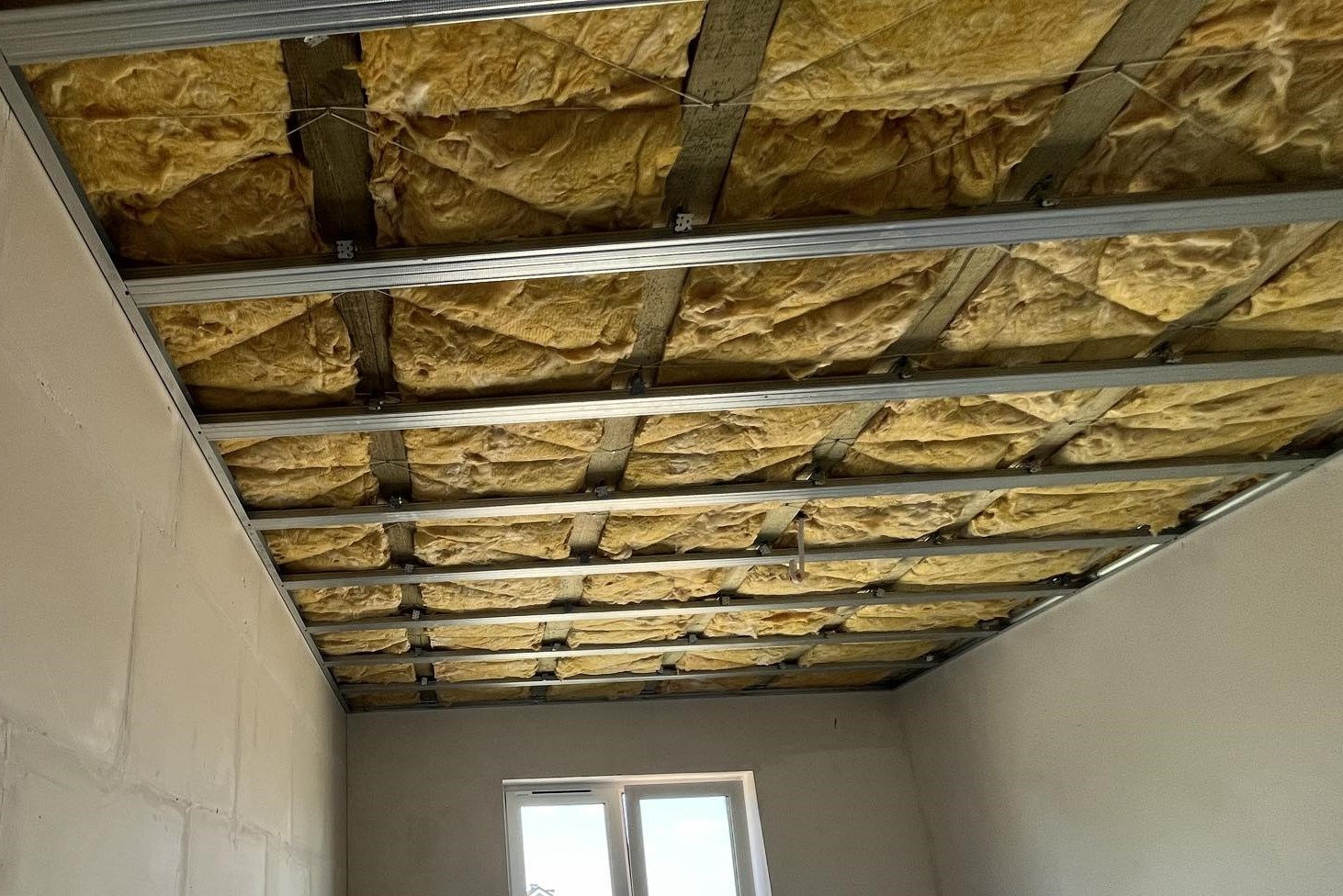 Montaż izolacji z wełny mineralnej w metalowym stelażu sufitu podwieszanego, widoczne fragmenty ścian i okno.