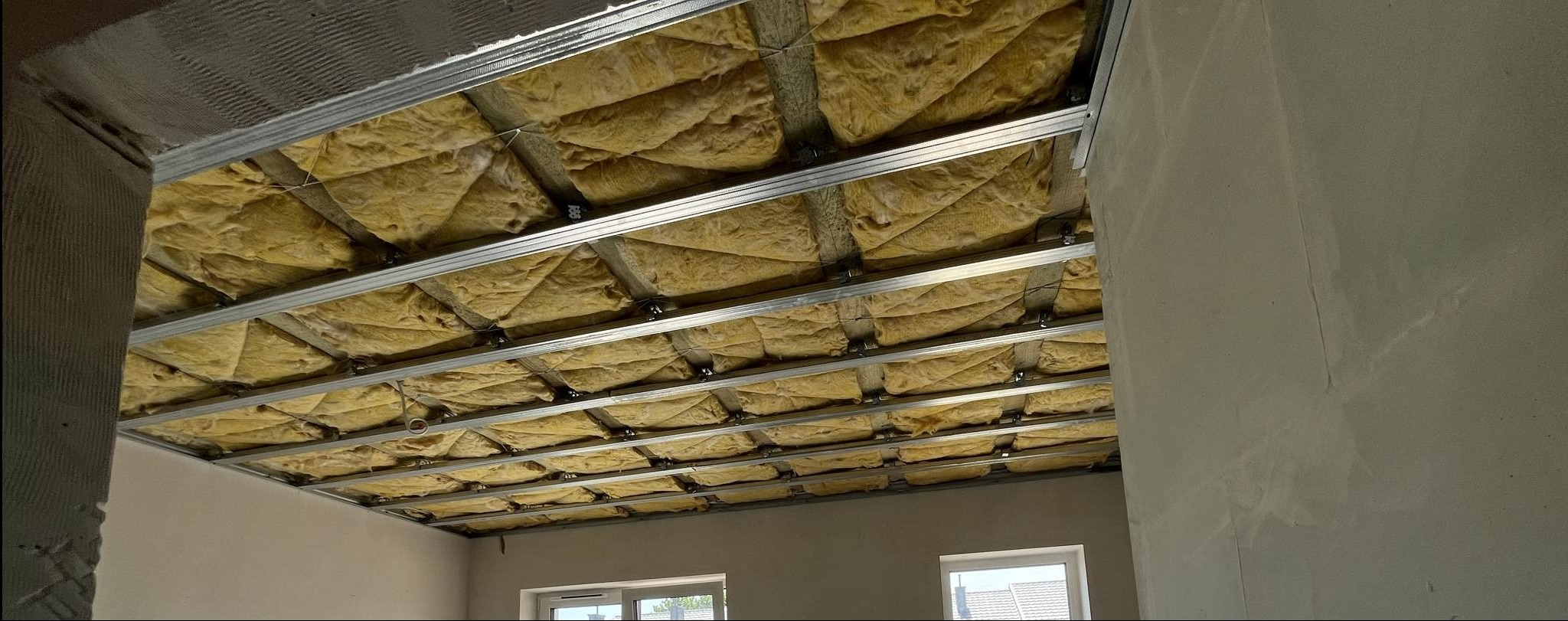 Montaż izolacji z wełny mineralnej w metalowym stelażu sufitu podwieszanego, widoczne okno w tle, ściany w stanie surowym.