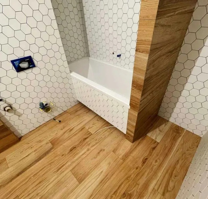 Wykończona łazienka z drewnopodobną podłogą, białą wanną obudowaną heksagonalnymi płytkami i ścianami wyłożonymi białymi płytkami heksagonalnymi z ciemnymi fugami, akcent z drewnopodobnych płytek...
