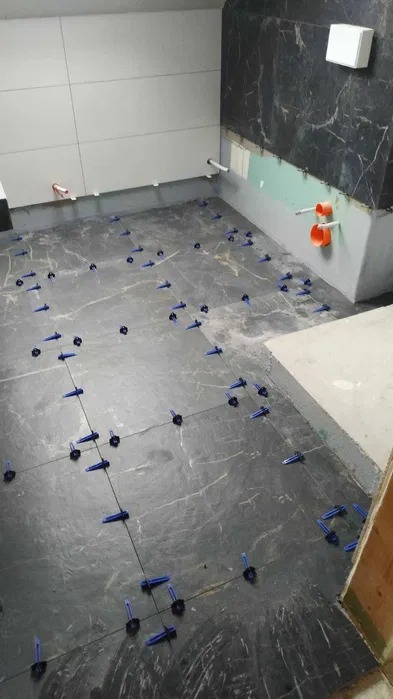 Układanie płytek podłogowych imitujących marmur z użyciem niebieskich klipsów poziomujących w trakcie remontu łazienki, widoczne fragmenty instalacji wodnej w ścianie.