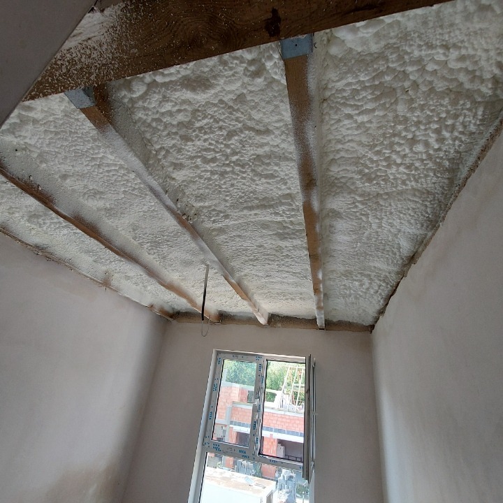 Poddasze z drewnianą konstrukcją stropu pokryte pianą izolacyjną, widoczne okno z widokiem na budowę.