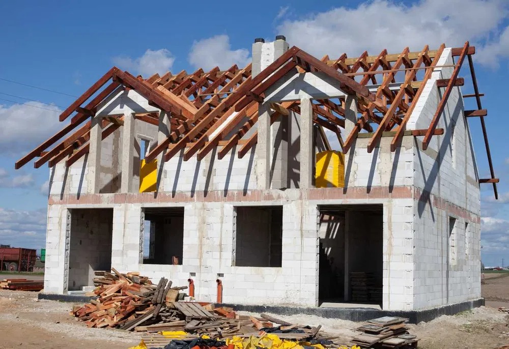 Surowy stan domu jednorodzinnego w trakcie budowy: ściany z białego bloczka, drewniana konstrukcja dachu bez pokrycia, żółte zabezpieczenia okien, stos drewna na pierwszym planie.