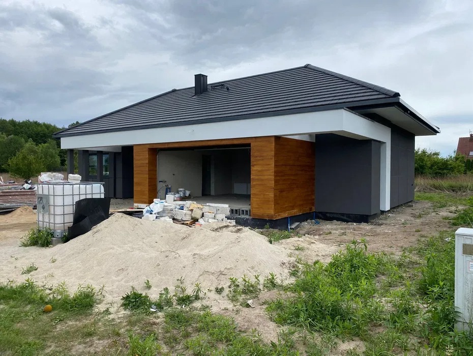 Nowoczesny dom jednorodzinny w trakcie budowy, z widoczną drewnianą elewacją garażu, szarą elewacją boczną, stertą piasku i materiałami budowlanymi przed budynkiem.