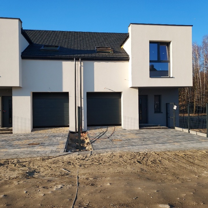 Nowoczesny szeregowiec z grafitowymi bramami garażowymi i kostką brukową przed wejściem, widoczne niedokończone prace ziemne.