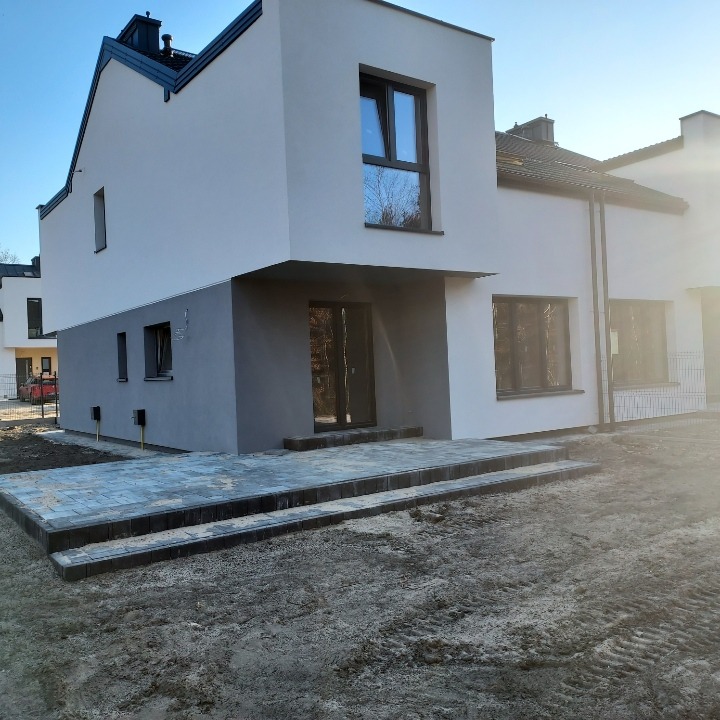 Nowoczesny dom jednorodzinny z dwukolorową elewacją (biała góra, szary dół) i wybrukowanym tarasem wejściowym, widoczne ślady opon na ziemi.