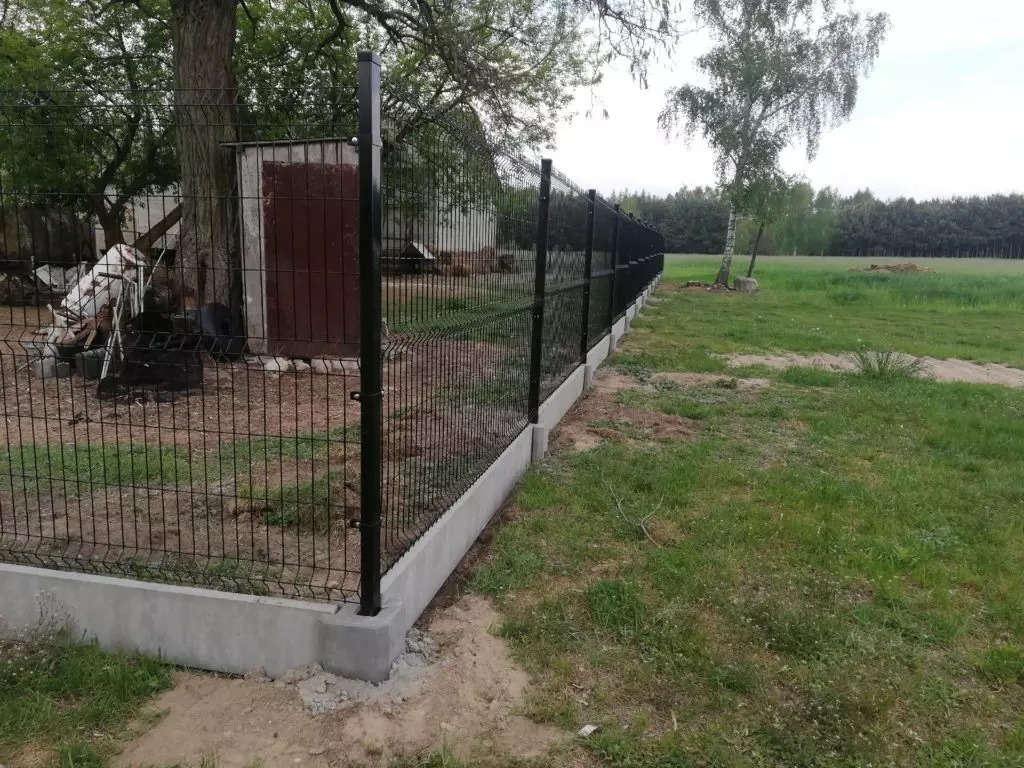 Długi, czarny panelowy system ogrodzeniowy na betonowej podmurówce, oddzielający posesję od łąki, z widocznym budynkiem gospodarczym w tle.