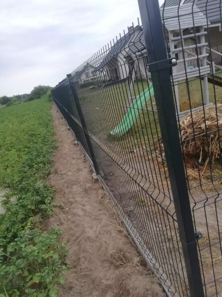 Długie, czarne ogrodzenie panelowe zamontowane na betonowej podmurówce, oddzielające ziemię uprawną od posesji z domem i placem zabaw dla dzieci w tle.