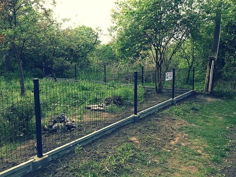 Czarny panel ogrodzeniowy zamontowany na betonowej podmurówce, widok z zewnątrz posesji, w tle zieleń i drzewa.