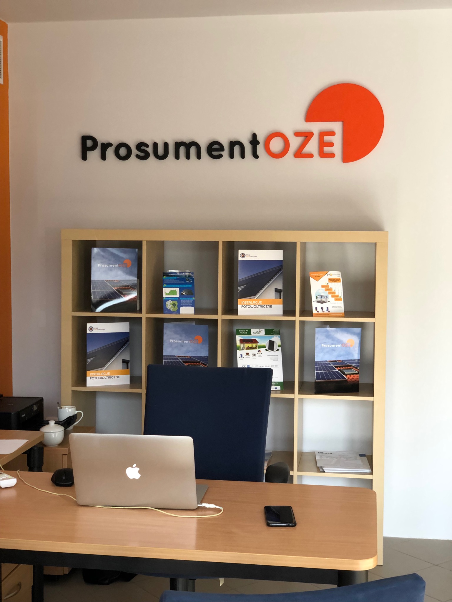 Biuro w Piekarach Śląskich z regałem wypełnionym broszurami informacyjnymi o instalacjach fotowoltaicznych i odnawialnych źródłach energii, laptop i telefon na biurku, logo firmy na ścianie.