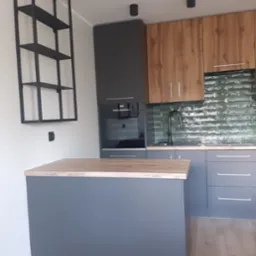 Szara kuchnia z drewnianymi elementami, wyspą i metalowym regałem na ścianie, zielone płytki na ścianie nad blatem.