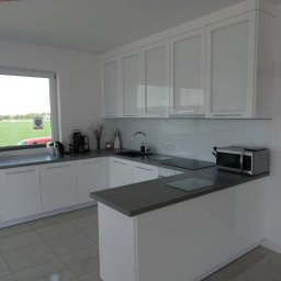 F.U.H.Armex Artur Piotrowski - Jasna kuchnia z białymi frontami szafek, szarym blatem i widokiem na zielone pole za oknem. Na blacie kuchennym ekspres do kawy, czajnik i mikrofalówka.