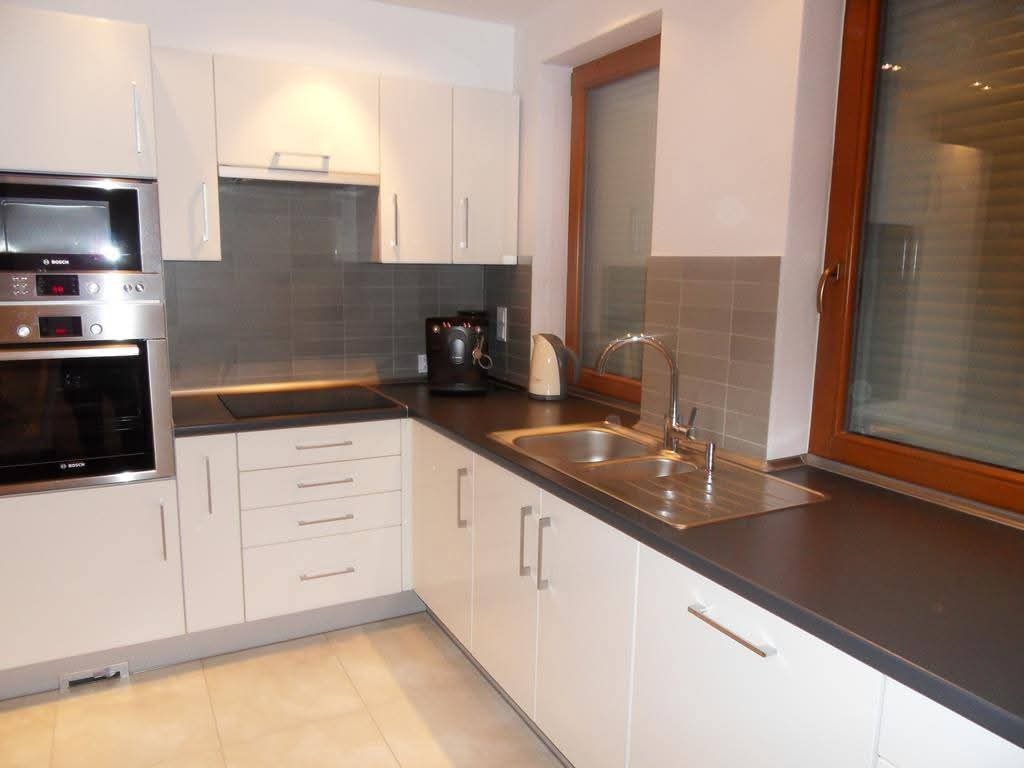 Jasna kuchnia z zabudową, sprzęt AGD w zabudowie, ciemny blat, okno z drewnianą ramą, płytki nad blatem w odcieniach szarości.