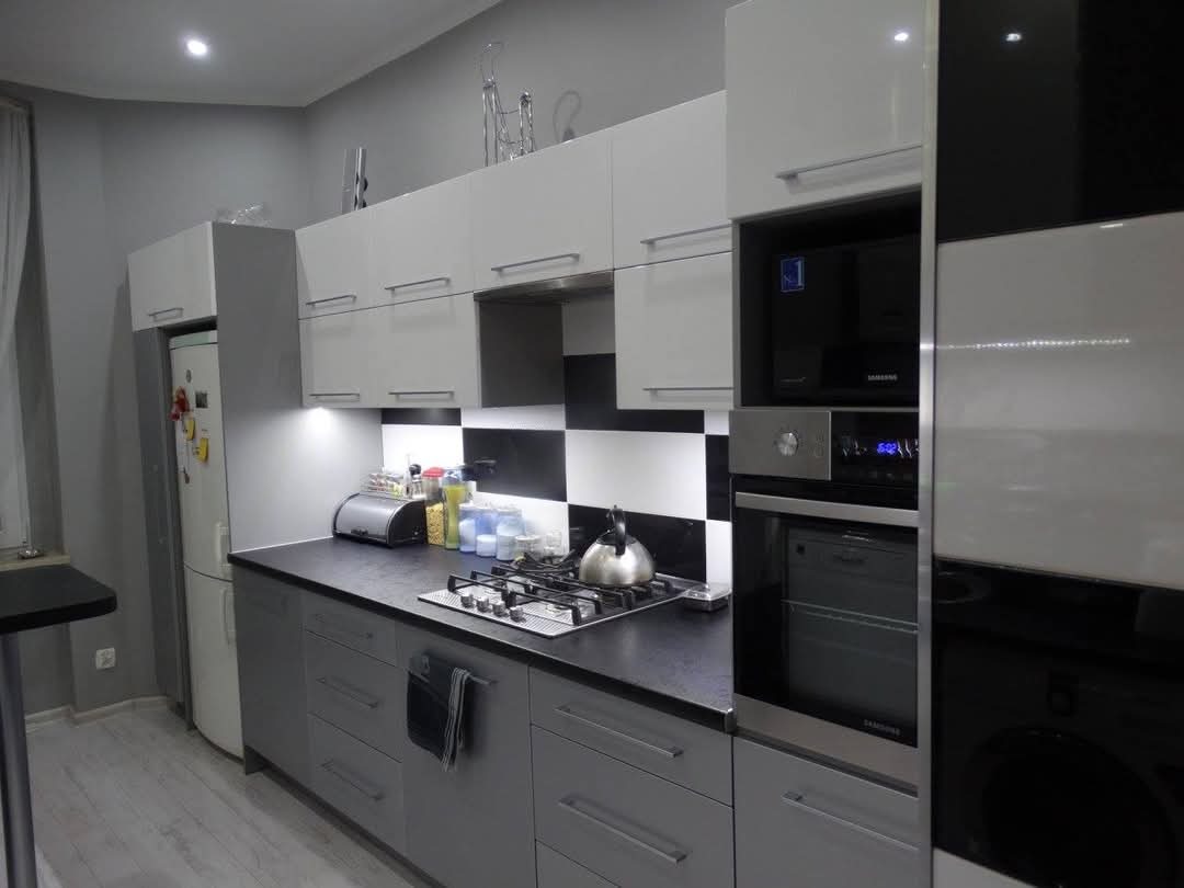 Nowoczesna kuchnia z szarymi szafkami, czarno-białymi płytkami na ścianie, zabudowanym piekarnikiem i mikrofalówką oraz czajnikiem na płycie gazowej