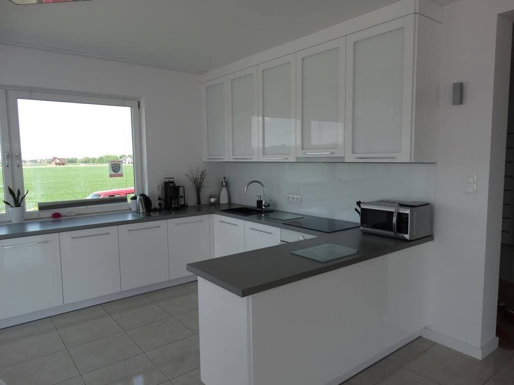 Jasna kuchnia z białymi frontami szafek, szarym blatem i widokiem na zielone pole za oknem. Na blacie kuchennym ekspres do kawy, czajnik i mikrofalówka.