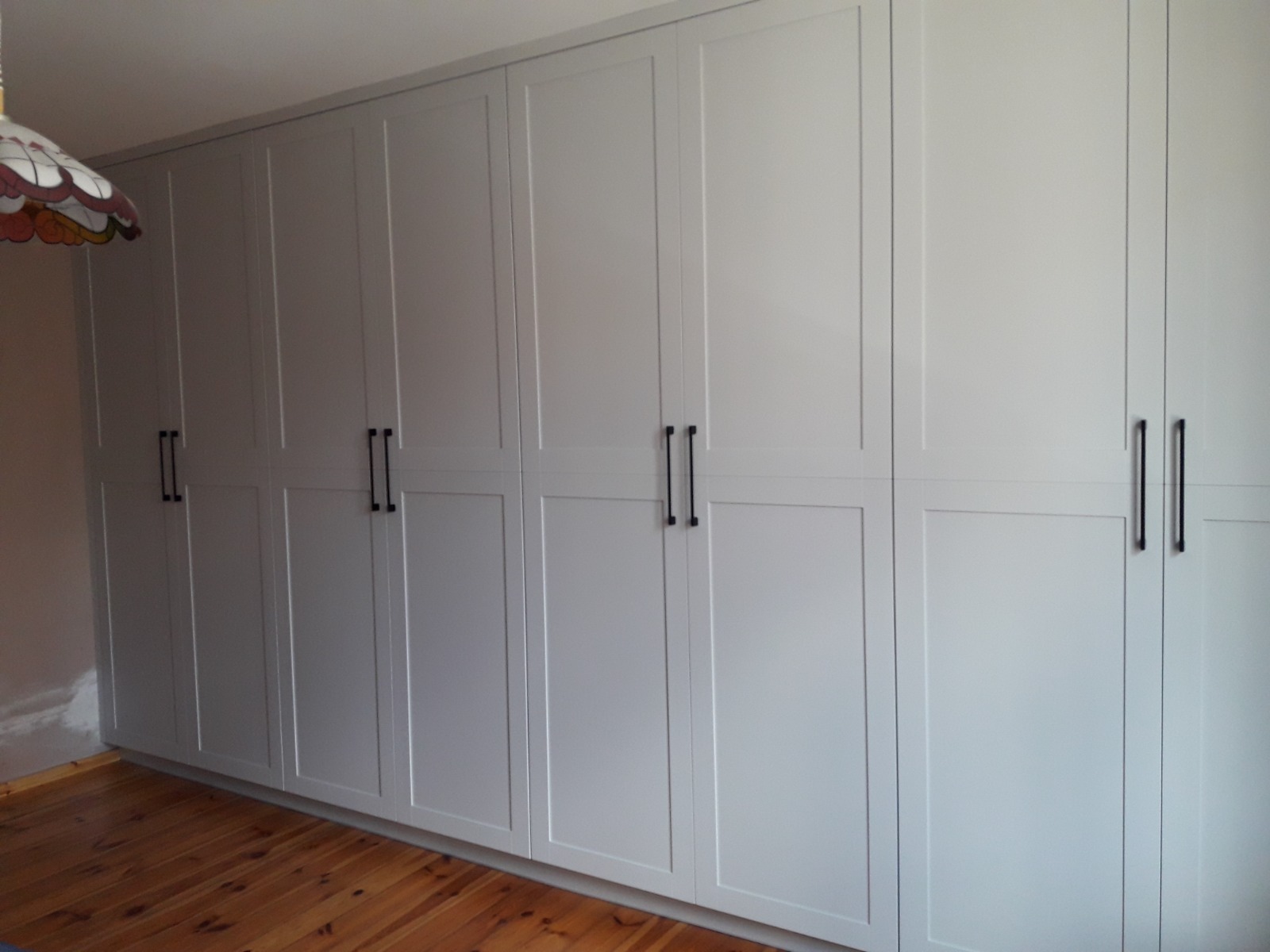 Długa, jasnoszara szafa wnękowa z czarnymi, pionowymi uchwytami, zajmująca całą ścianę, na tle drewnianej podłogi i fragmentu lampy w stylu vintage.