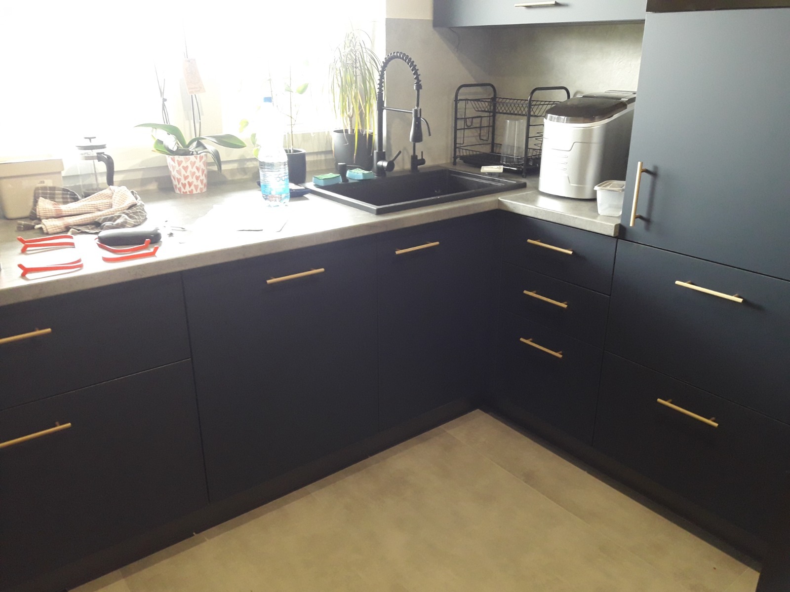 Ciemna, matowa kuchnia narożna z betonowym blatem, złotymi uchwytami, czarnym zlewem i baterią, oraz różnymi akcesoriami kuchennymi na blacie pod oknem.