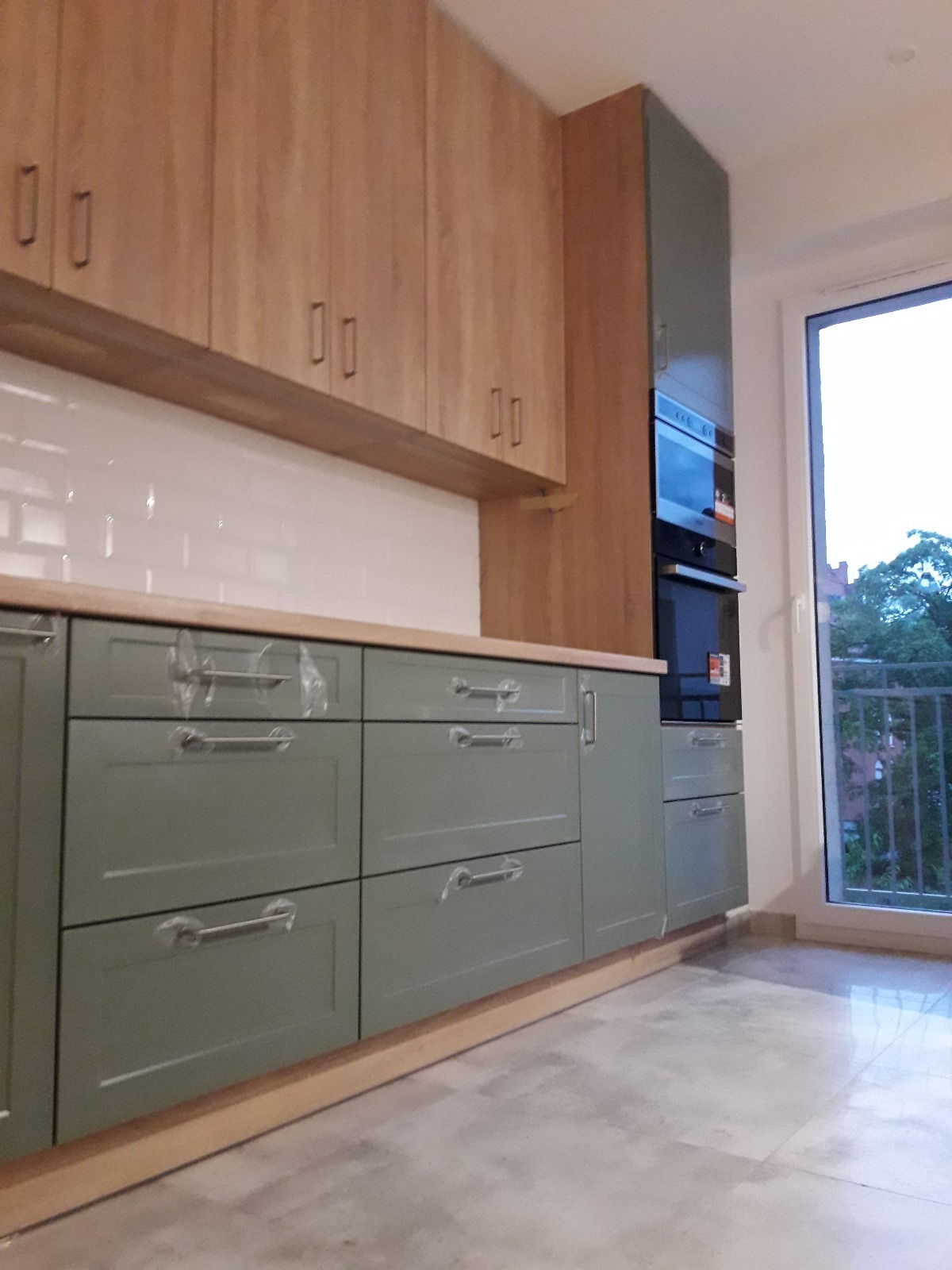 Nowoczesna kuchnia z drewnianymi szafkami górnymi i zielonymi szafkami dolnymi z srebrnymi uchwytami, z piekarnikiem w zabudowie i widokiem na zewnątrz przez okno.