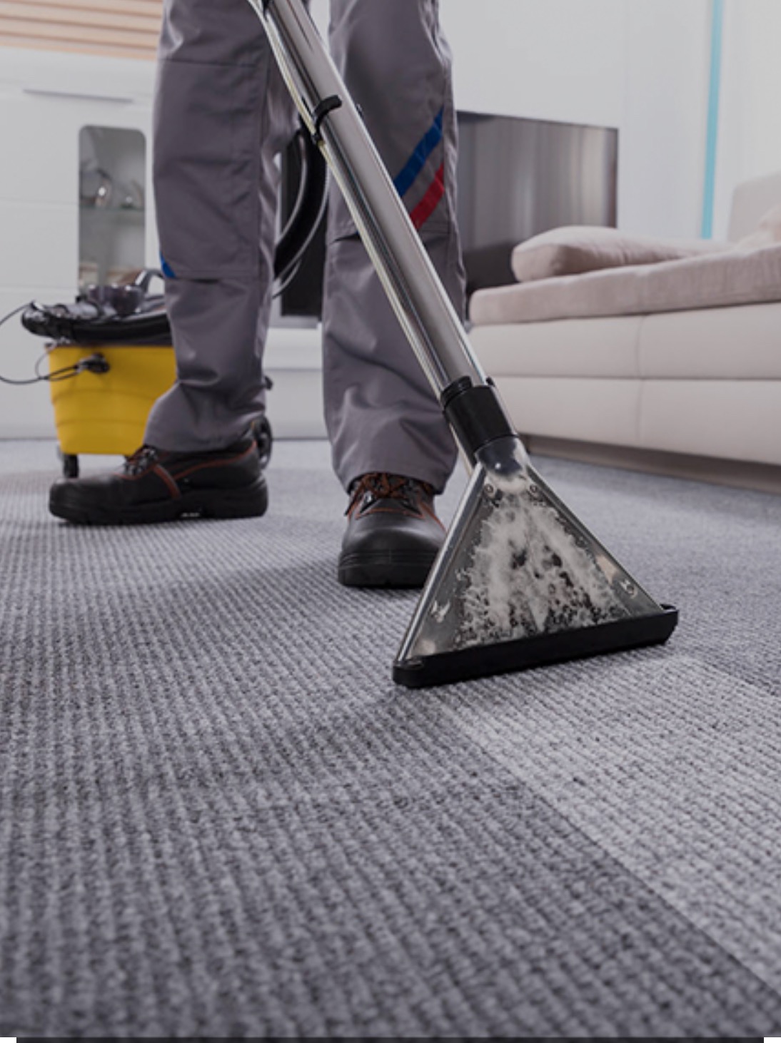 Czyszczenie szarego dywanu odkurzaczem piorącym; widoczna brudna woda w przezroczystej ssawce, pracownik w szarym kombinezonie i czarnych butach.