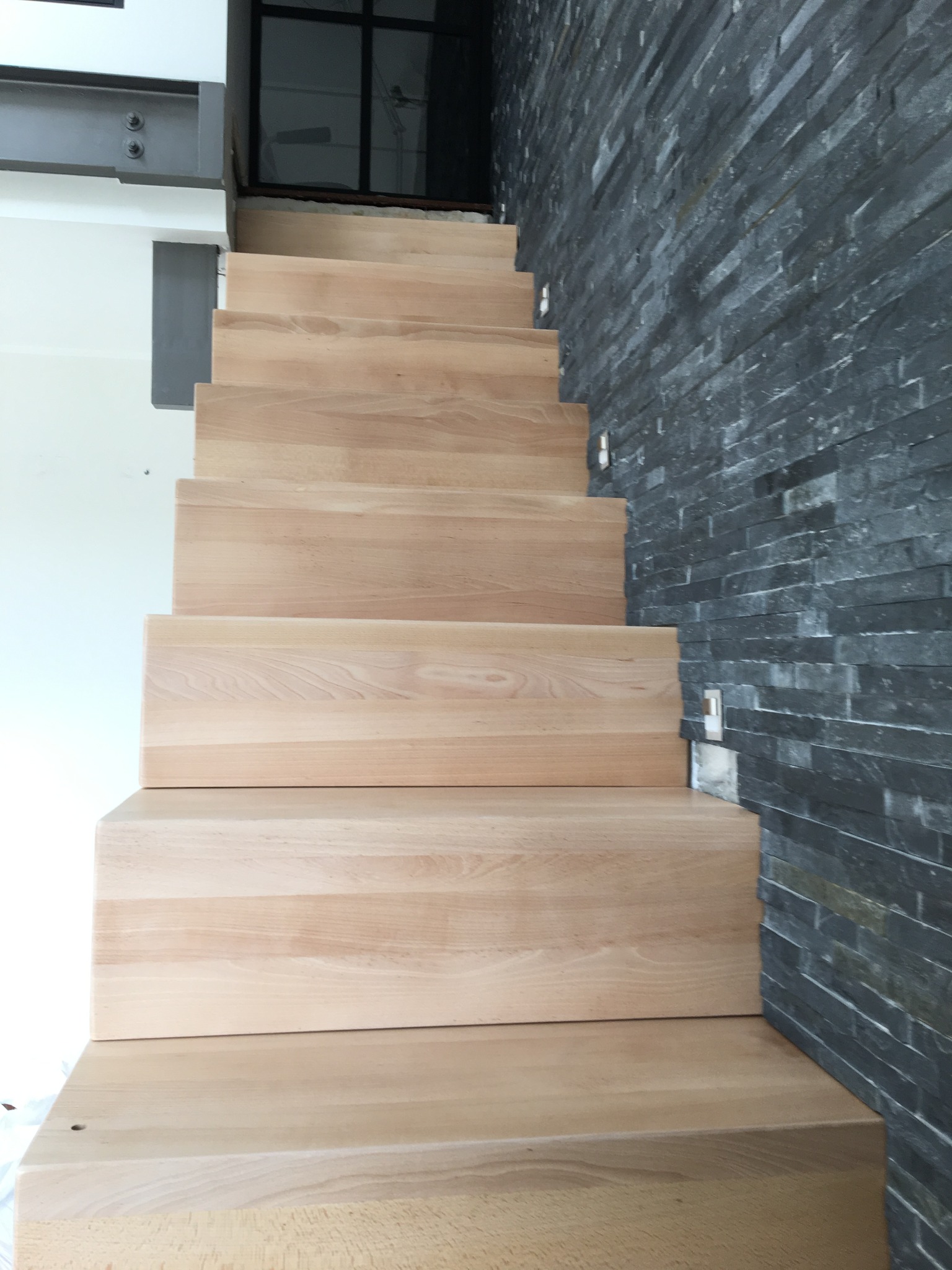 Nowoczesne schody drewniane z jasnego drewna, widok z dołu, z ciemną ścianą z kamienia łupkowego po prawej stronie.