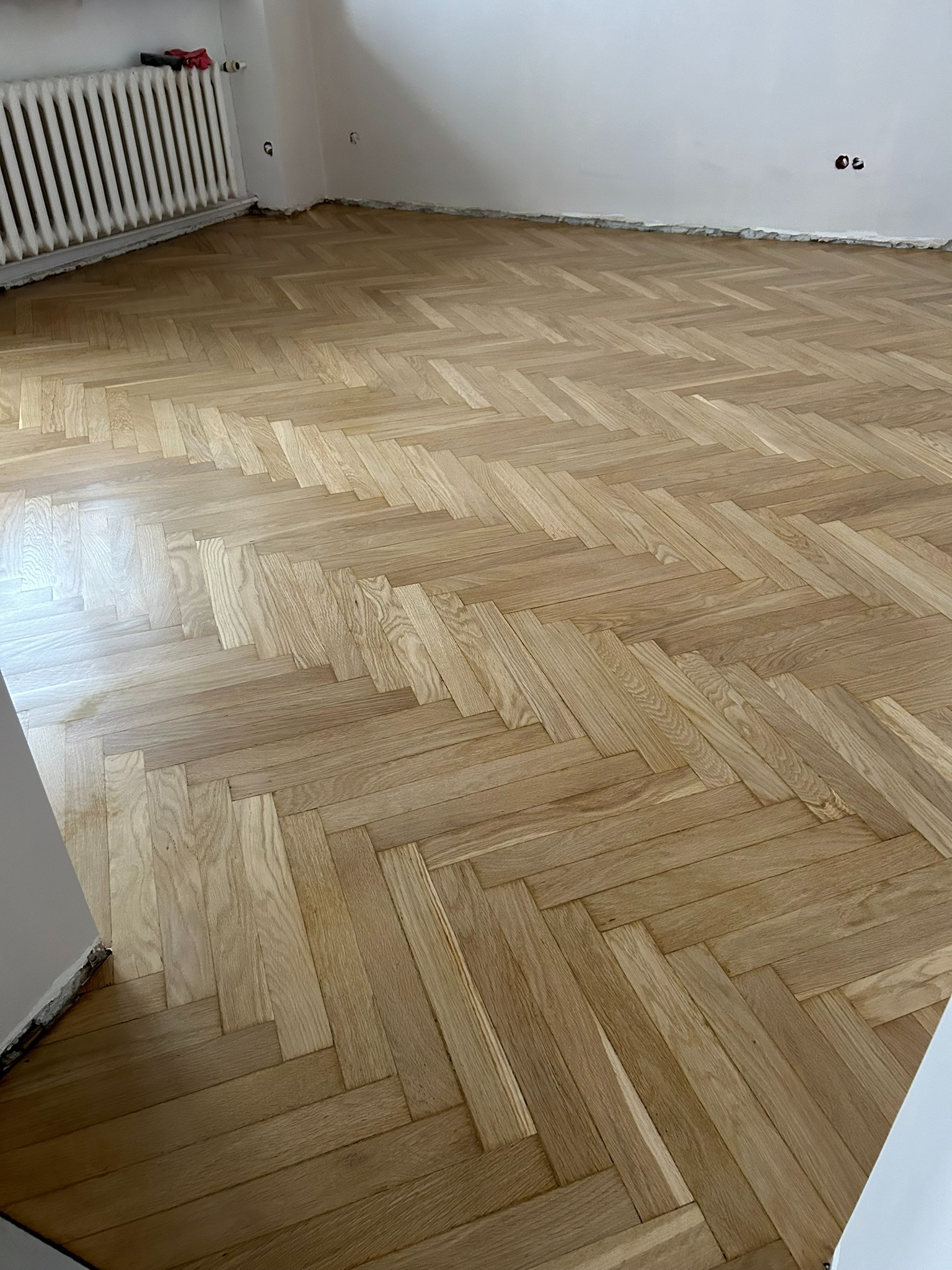 Parkiet ułożony w jodełkę po renowacji, widoczny grzejnik i ślady po listwach przypodłogowych na ścianie.
