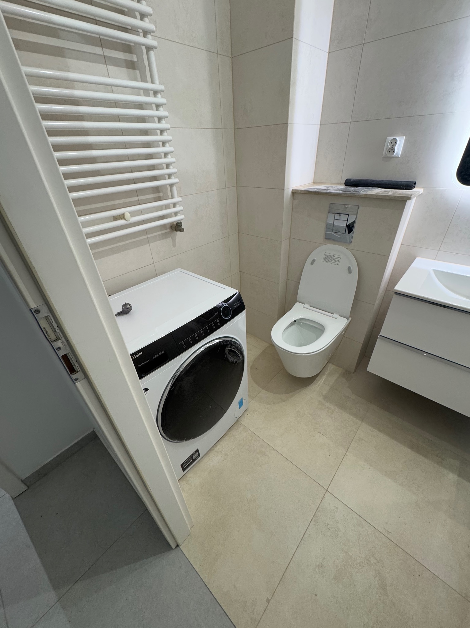 Jasna łazienka po remoncie z pralką Haier, toaletą podwieszaną i grzejnikiem drabinkowym na ścianie z płytek w odcieniach beżu.