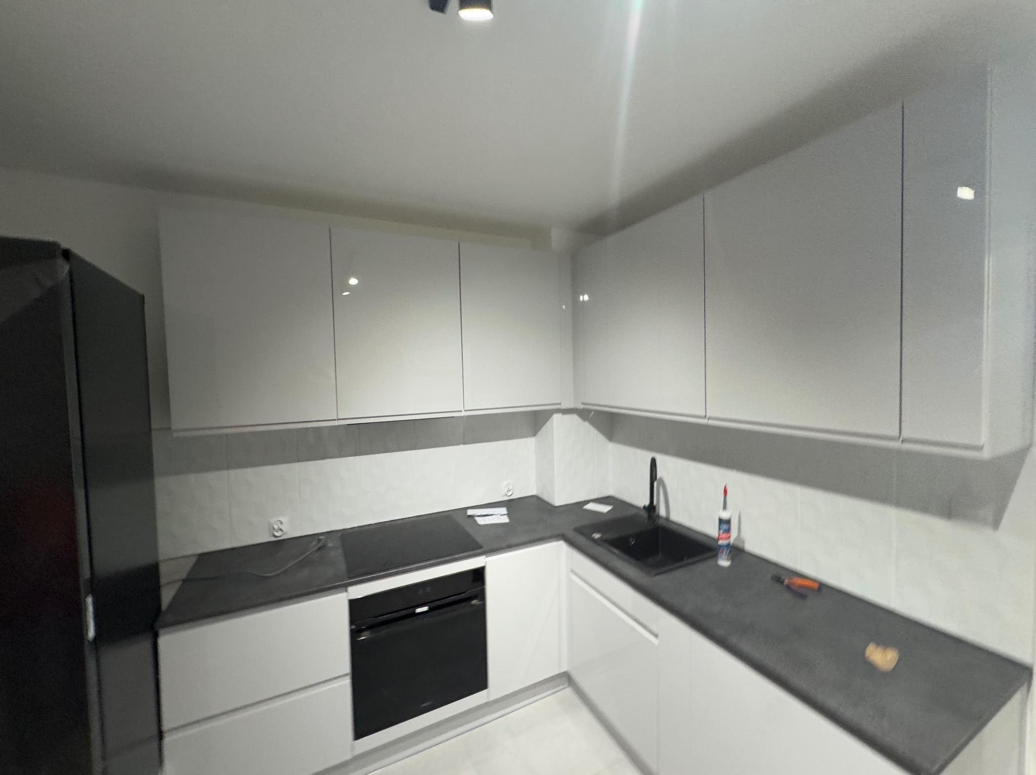 Nowoczesna kuchnia z białymi szafkami, szarym blatem i czarnym sprzętem AGD, w tym piekarnikiem i zlewem z baterią. Na blacie widoczne akcesoria montażowe i silikon. Ściany wyłożone białymi...