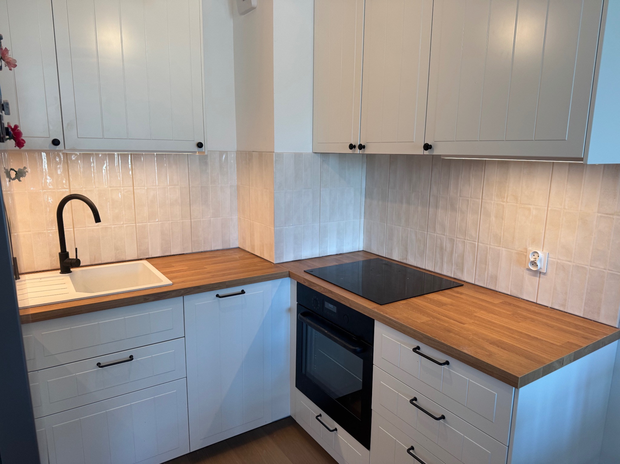 Biała kuchnia z drewnianym blatem, czarną armaturą i płytą indukcyjną, oraz płytkami imitującymi cegłę na ścianie.