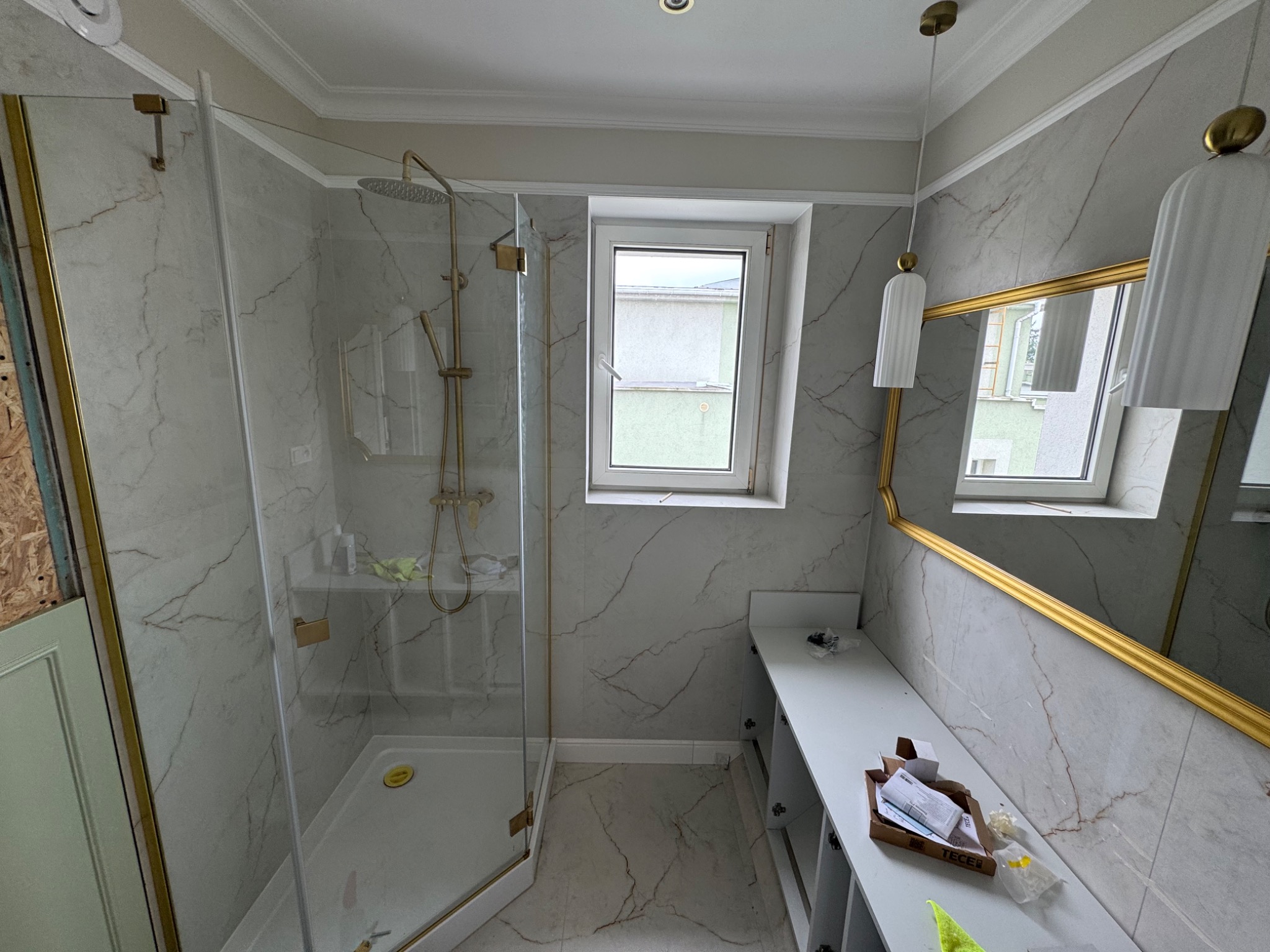 Wykończona łazienka z kabiną prysznicową typu walk-in, marmurowymi płytkami, złotymi akcentami i oświetleniem. Widoczne elementy montażowe.