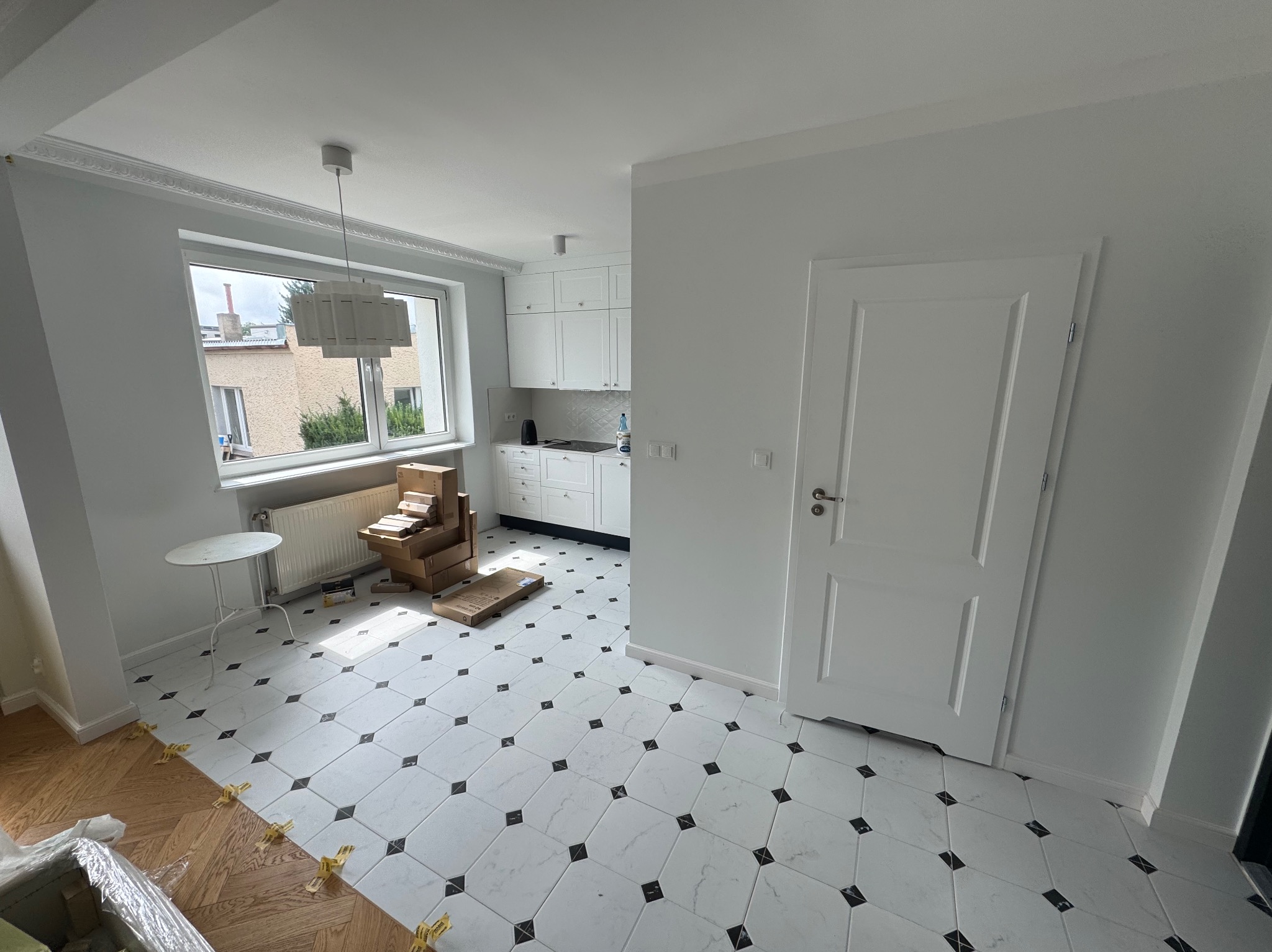 Jasne wnętrze po remoncie: białe ściany, nowa podłoga w biało-czarne romby, biała kuchnia w tle. Część podłogi jeszcze nieukończona, widoczne płytki i akcesoria.