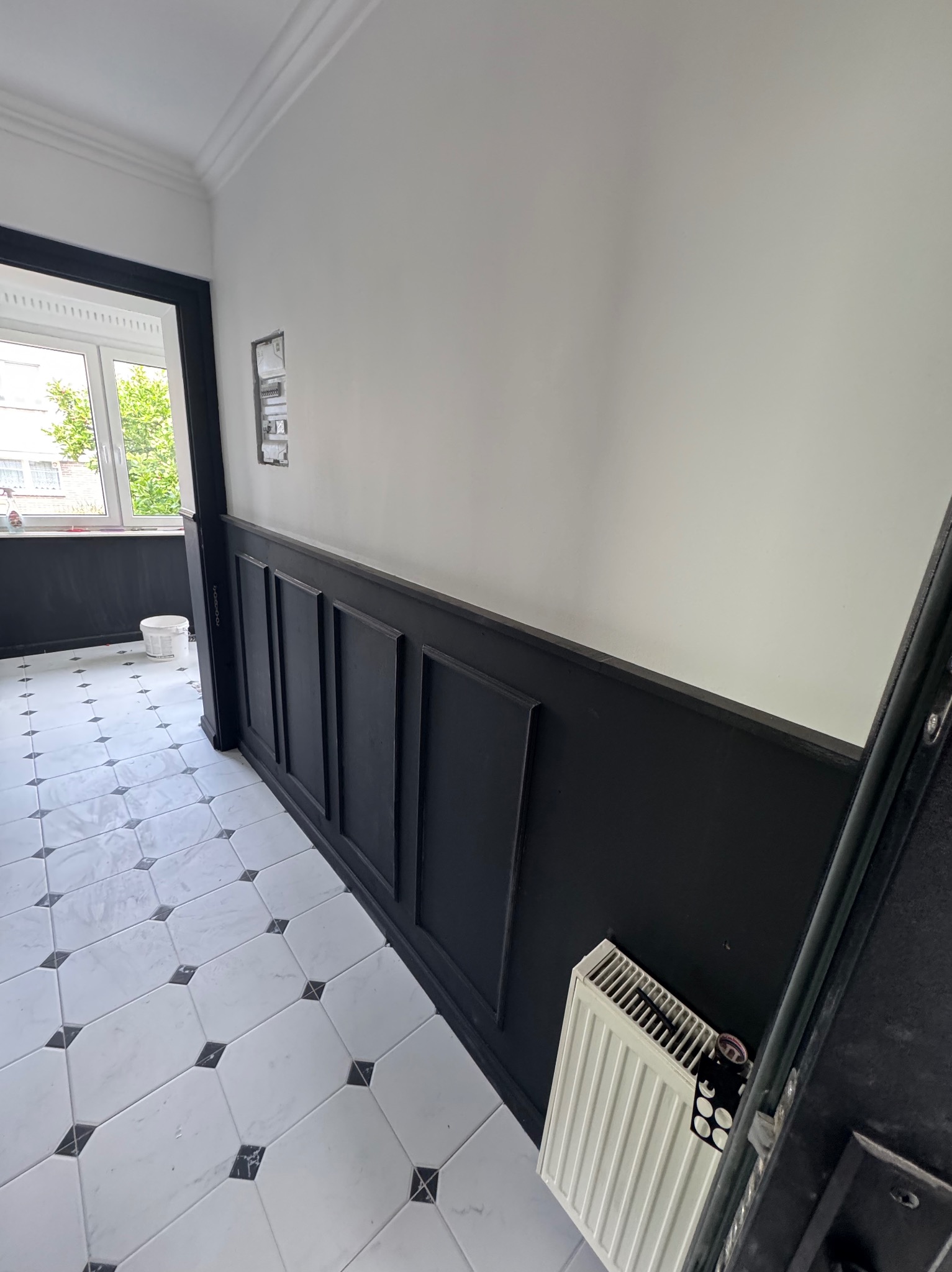 Wykończony korytarz z panelami ściennymi w kolorze czarnym, białą górną częścią ścian i marmurową podłogą w biało-czarne wzory. Widoczny grzejnik i fragment okna.