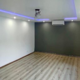 Usługi remontowo - budowlane DARBUD - Nowoczesne wnętrze po remoncie: jasna podłoga z paneli, białe ściany kontrastujące z szarą ścianą, sufit z oświetleniem LED i lampą wiszącą, klimatyzator.
