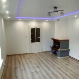 Usługi remontowo - budowlane DARBUD - Jasne panele podłogowe w pokoju z białymi ścianami, podwieszanym sufitem z oświetleniem LED i kominkiem w rogu