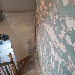 Usługi remontowo - budowlane DARBUD - Klatka schodowa w trakcie przygotowywania ścian do malowania, widoczne ślady po zdrapywaniu starej farby, surowe schody i fragment balustrady.