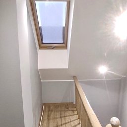 Usługi remontowo - budowlane DARBUD - Klatka schodowa z drewnianymi panelami podłogowymi, pomalowana na szaro ściana i drewniana balustrada, okno dachowe w górnej części klatki schodowej.