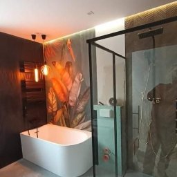 Usługi remontowo - budowlane DARBUD - Nowoczesna łazienka z białą wanną, kabiną prysznicową z czarną ramą, grzejnikiem drabinkowym, i tapetą w tropikalne liście, oświetlona wiszącymi lampami.