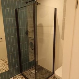 Usługi remontowo - budowlane DARBUD - Nowoczesna kabina prysznicowa z czarnymi profilami i szklanymi ściankami, widoczna na tle turkusowych płytek ułożonych pionowo oraz wzorzystej podłogi. W tle pralka i fragment toalety.