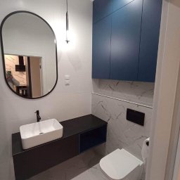 Usługi remontowo - budowlane DARBUD - Nowoczesna łazienka z owalnym lustrem w czarnej ramie, wiszącą szafką z umywalką, podwieszaną toaletą i szafkami w kolorze ciemnego błękitu, wykończona płytkami imitującymi biały marmur.