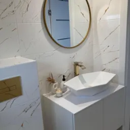 Nowoczesna łazienka z białymi płytkami imitującymi marmur ze złotymi żyłami, okrągłe lustro w złotej ramie, biała szafka z umywalką o geometrycznym kształcie, złota bateria, dekoracje: wazonik...
