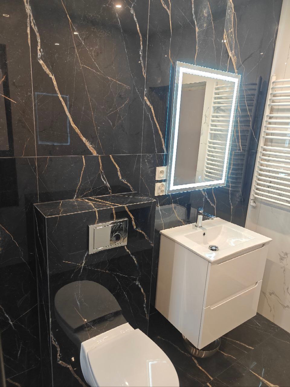 Nowoczesna łazienka z czarnymi marmurowymi płytkami, podświetlanym lustrem LED, białą umywalką z szafką i wiszącą toaletą.