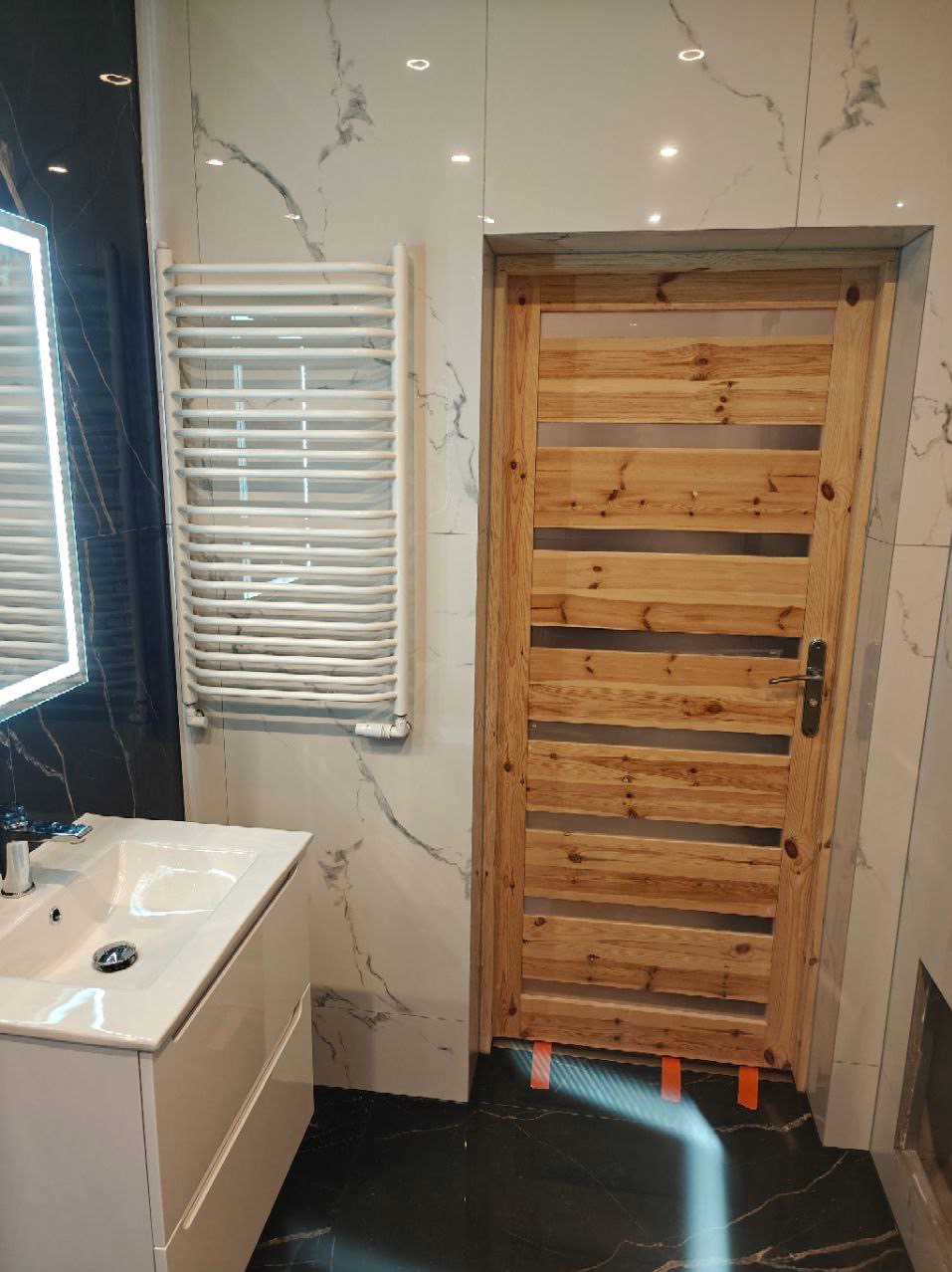 Nowoczesna łazienka z białym umywalką na szafce, grzejnikiem drabinkowym i drewnianymi drzwiami z poziomymi przeszkleniami, wykończona płytkami imitującymi marmur.