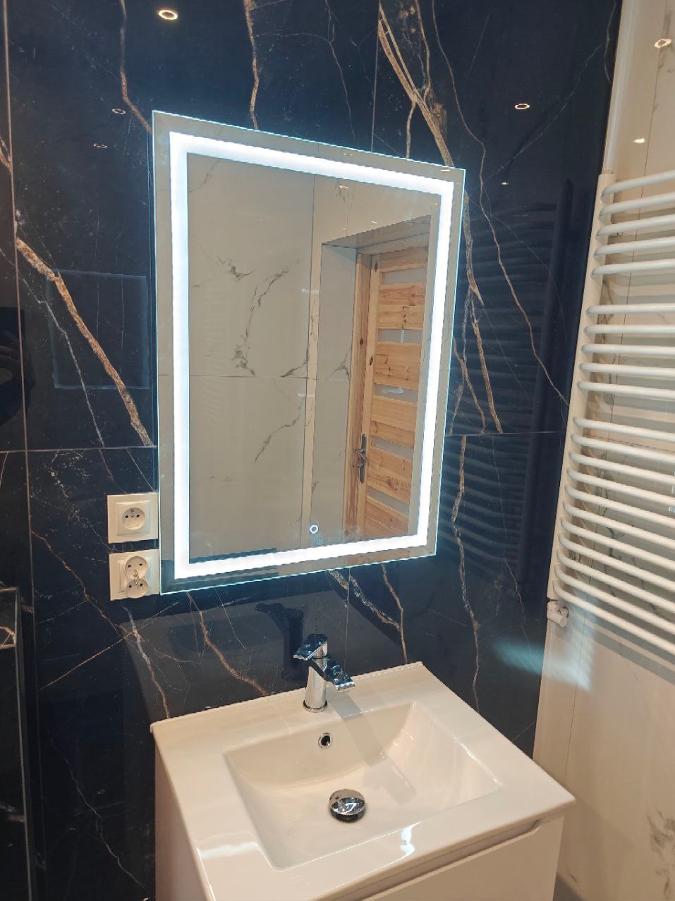 Nowoczesna łazienka z czarnymi marmurkowymi płytkami, podświetlanym lustrem LED, białą umywalką i grzejnikiem drabinkowym.