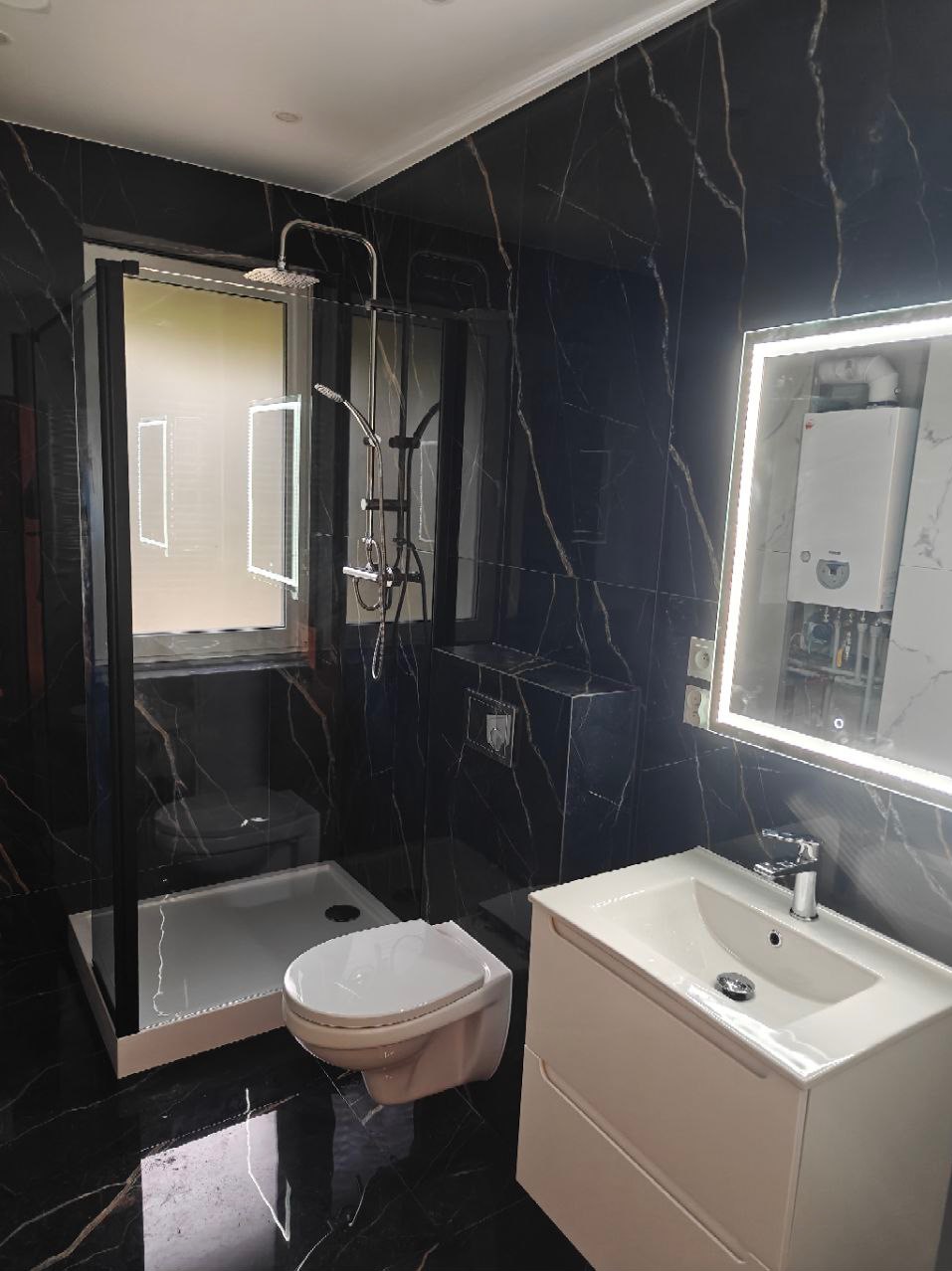 Nowoczesna łazienka z czarnymi marmurkowymi płytkami, kabiną prysznicową typu walk-in z czarną ramą, podwieszaną toaletą i białą szafką z umywalką podświetlaną lustrem.