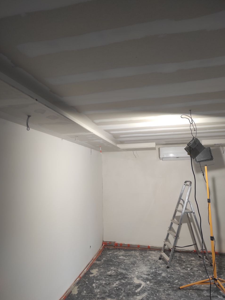 Pokój w trakcie wykańczania: sufit podwieszany z płyt gipsowo-kartonowych, ściany przygotowane do malowania, drabina aluminiowa i lampa budowlana na trójnogu oświetlają wnętrze.