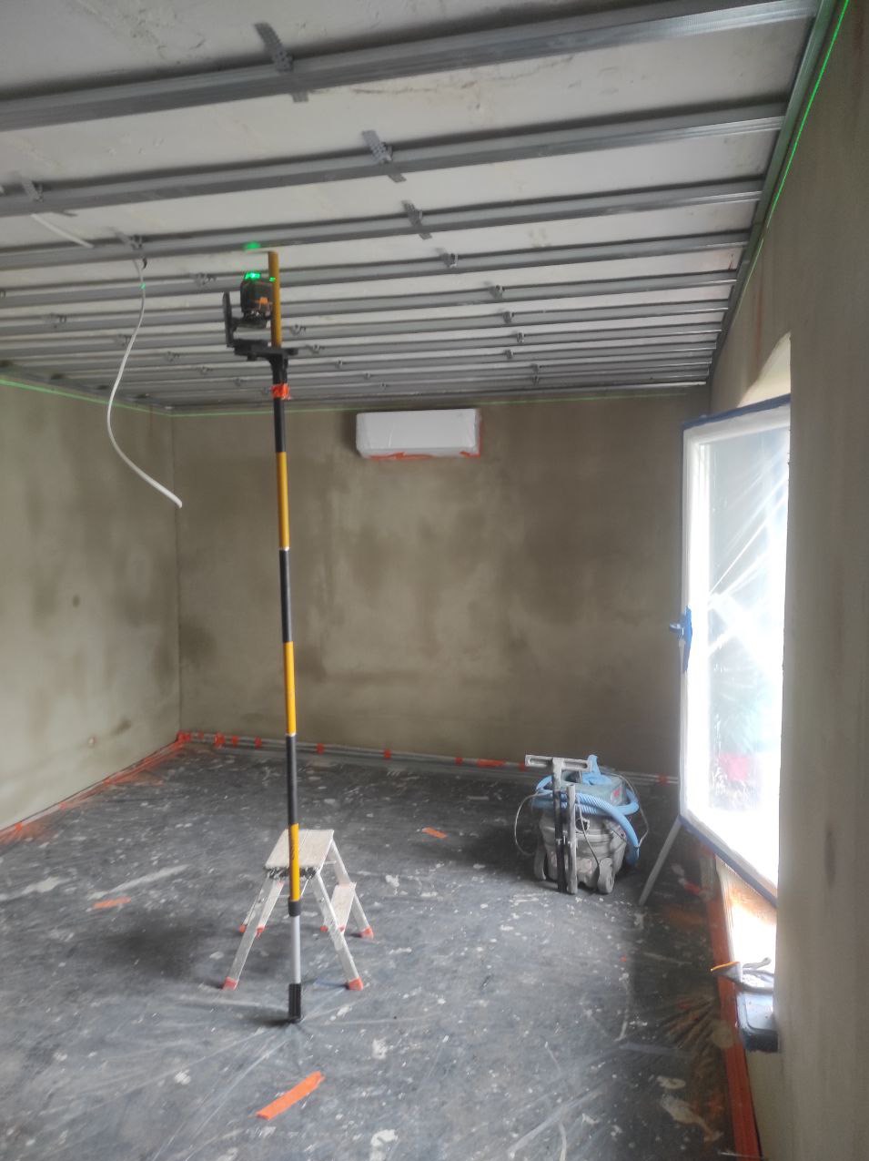 Pokój w trakcie remontu, widoczny laser krzyżowy na statywie, szlifierka do gładzi, profile sufitowe, zabezpieczone okno folią i ściany po gruntowaniu.