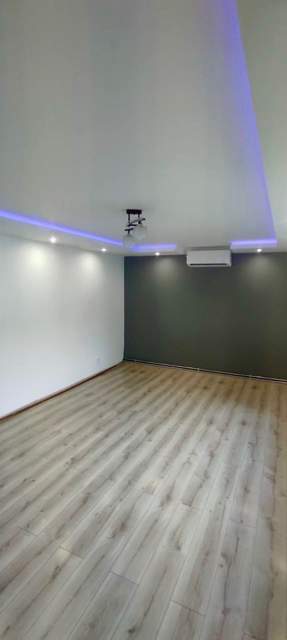 Nowoczesne wnętrze po remoncie: jasna podłoga z paneli, białe ściany kontrastujące z szarą ścianą, sufit z oświetleniem LED i lampą wiszącą, klimatyzator.