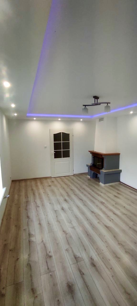 Jasne panele podłogowe w pokoju z białymi ścianami, podwieszanym sufitem z oświetleniem LED i kominkiem w rogu