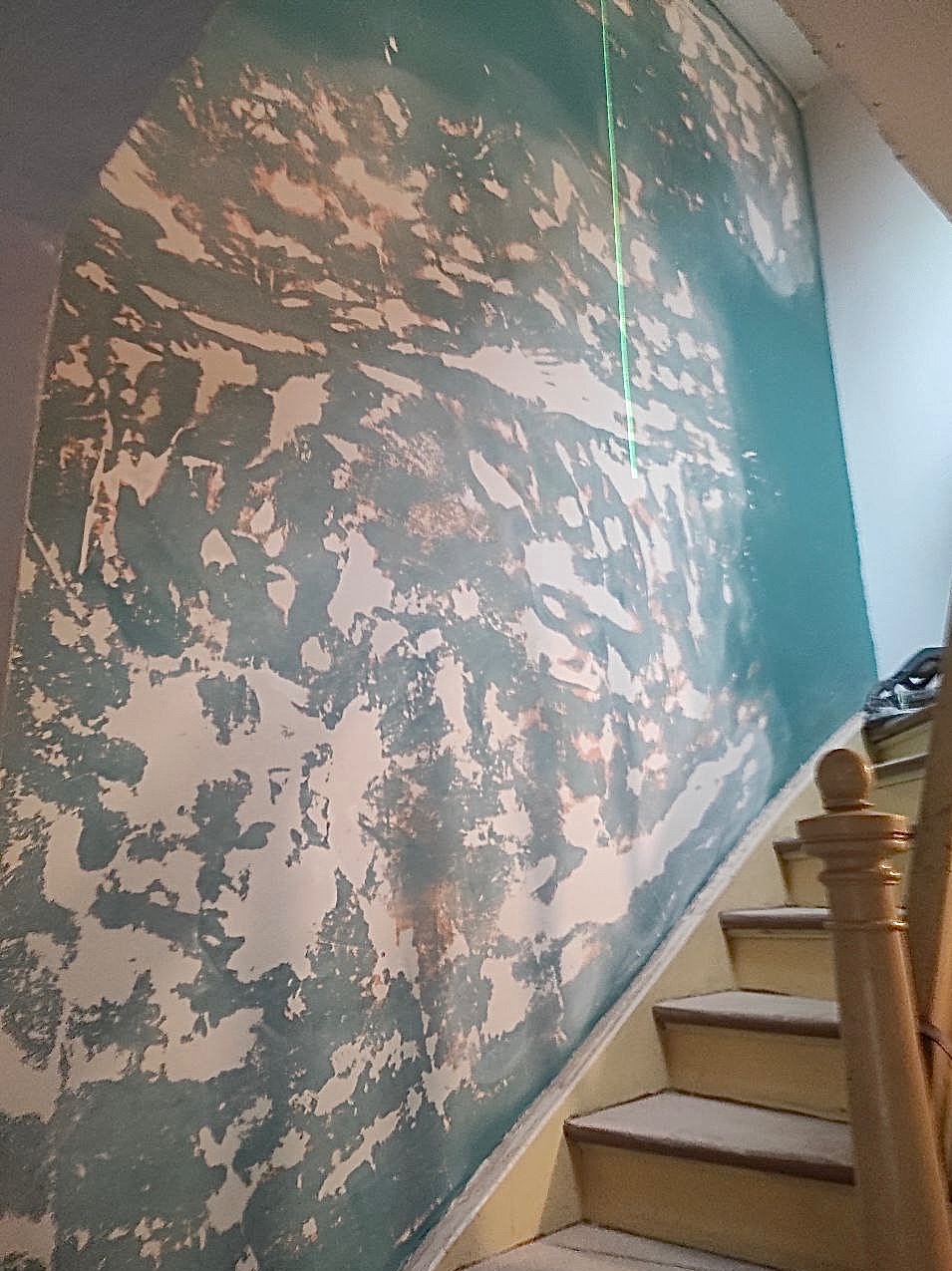 Ściana w trakcie malowania z widocznymi warstwami farby i zieloną linią lasera, obok schody z drewnianą balustradą.