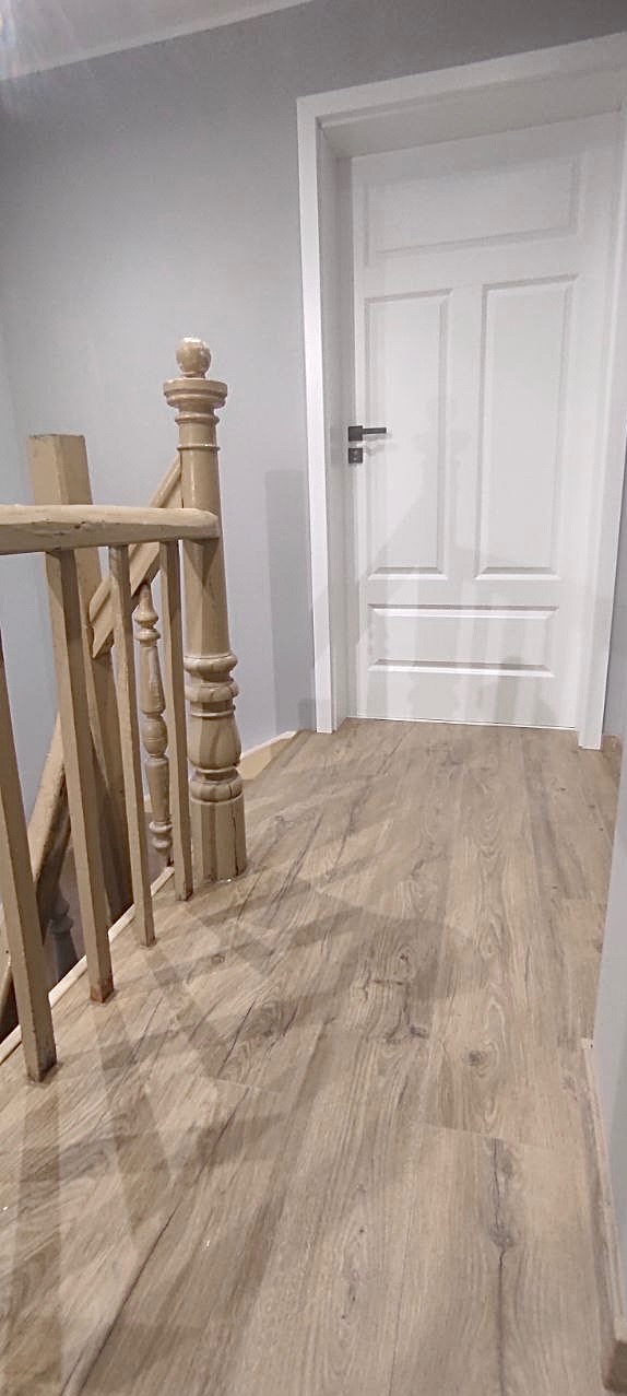 Jasne panele podłogowe z drewnianym wzorem ułożone w korytarzu w Prusicach, widoczne schody z beżową balustradą i białe drzwi.