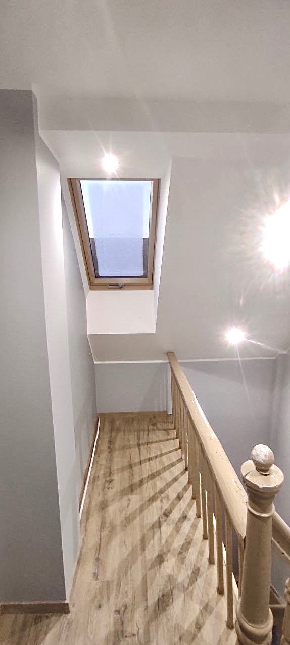 Klatka schodowa z drewnianymi panelami podłogowymi, pomalowana na szaro ściana i drewniana balustrada, okno dachowe w górnej części klatki schodowej.